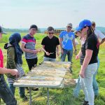 University-age students stand outdoors around a table with plants on it during an agronomy-focused field trip.