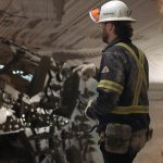 A miner clad in coveralls, wearing gloves and a hard hat, is gesturing with his hands underground in a potash mine.