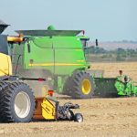 Two combines, a yellow one and a green one, kick up dust as they harvest side by side.