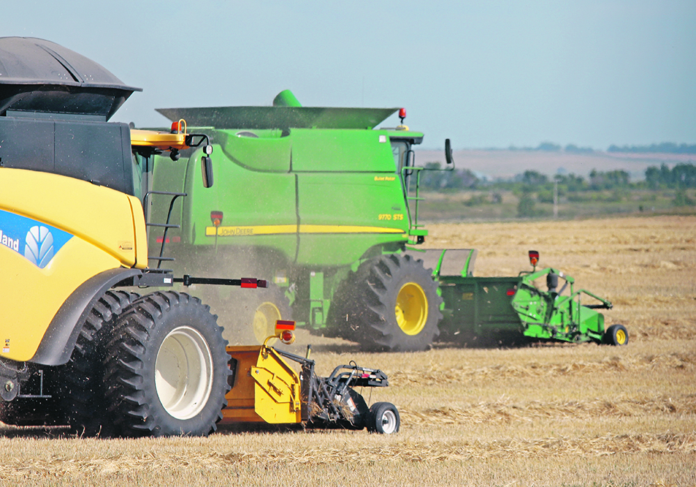 Two combines, a yellow one and a green one, kick up dust as they harvest side by side.