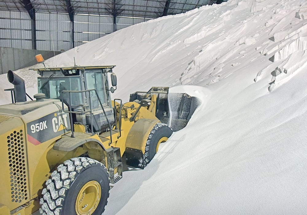 A wheel loader scoops up a bucket full of urea fertilizer from a massive pile in an indoor warehouse.