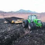 A large, green, 8-wheeled tractor pulls a tiller through a harvested peat bog as part of restoration efforts.
