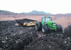 A large, green, 8-wheeled tractor pulls a tiller through a harvested peat bog as part of restoration efforts.