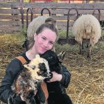 Kamden Bartman smiles for the camera holding a young member of her flock while kneeling in a pen.