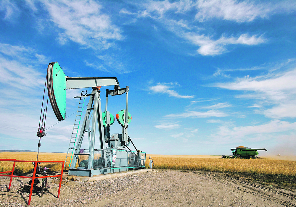 An oilfield pumpjack sits on a gravel pad in the midst of a ripe wheat field with a combine passing by in the background.