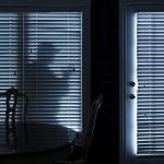 A stock photo of a shadow, possibly a robber, visible through the blinds of a darkened room at night.