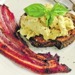 Scrambled eggs on top of a piece of rye bread toast with garnish and several strips of bacon on a plate.