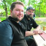 Two men, James Kinley in the foreground and Reuben Kolk in the background, sit at a picnic table in a park.
