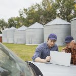 An older farmer and a young farmer look at a laptop resting on the hood of a vehicle with grain bins in the background.