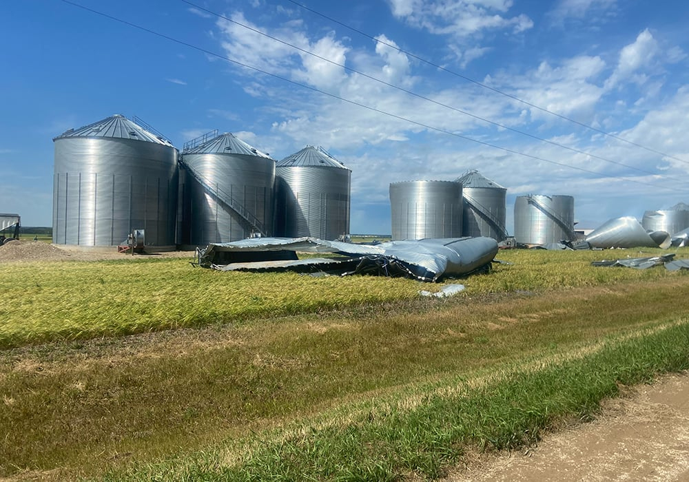 A flattened metal grain bin is on the ground in front of six others that remain standing in the aftermath of a atorm.