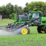 An agricultural high-clearance sprayer is seen spraying a lush, green crop.