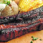 A juicy, barbecued rib sits on a wooden cutting board with a baked potato topped with sour cream and a couple cobs of corn in the background.