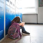 A young girl sits slumped against the lockers in a school with her backpack beside her.