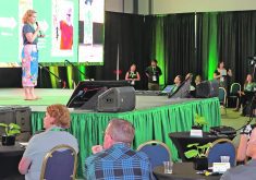 A woman with a microphone is on a stage addressing a group of people seated at tables in a large hall.