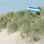 A person in a bathing suit sits under a blue and white umbrella amid beach grass on a sandy beach dune.