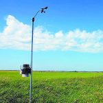 A small weather station is mounted on a pole in front of a farmer's field.