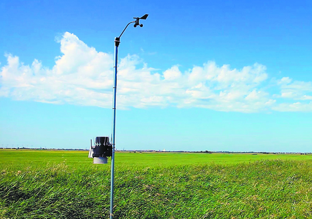 A small weather station is mounted on a pole in front of a farmer's field.