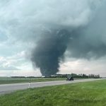 A vehicle drives by on a highway in the foreground while a dark and massive tornado spins in the background.