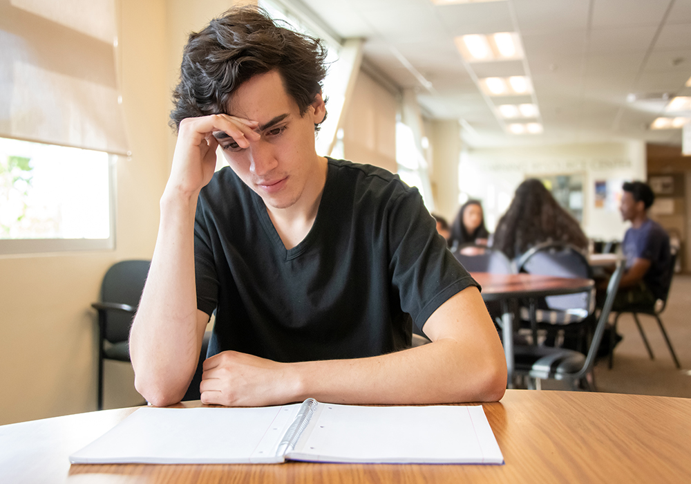 A college student sits alone in a library looking over his notebook.