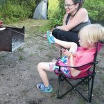 A woman and a child roast weiners on sticks over a fire in a metal campfire box.