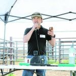 A man in a baseball cap speaks into a microphone with cattle pens in the background