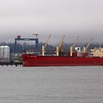 A red cargo ship at port in Vancouver with large metal bins on the dock to the left.