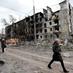 A woman walks down a street in a bombed out Ukraining town, a destroyed 5-storey appartment complex is visible in the background.