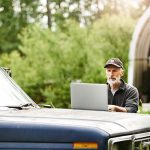 A farmer looks at a laptop computer that is sitting on the hood of an old truck.