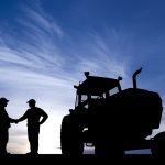 Two farmers, silhouetted against the setting sun, shake hands next to a tractor.