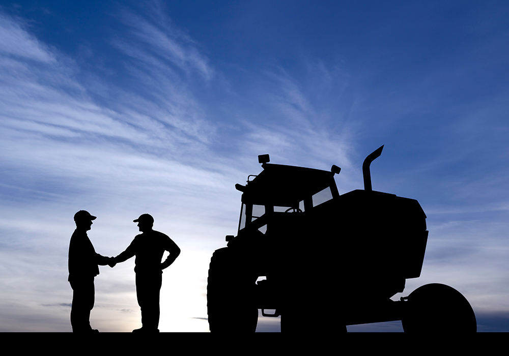 Two farmers, silhouetted against the setting sun, shake hands next to a tractor.