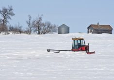 An old farmyard in winter. A swather sits in deep snow with an old shed and metal grain bin in the background.