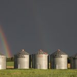 A rainbow arcs to the ground against a dark sky behind 5 round, metal grain bins.