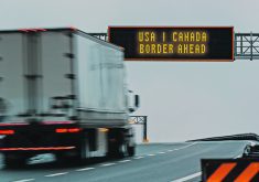 A semi truck moves down a highway about to pass beneath a sign that reads, " USA | Canada Border Ahead."