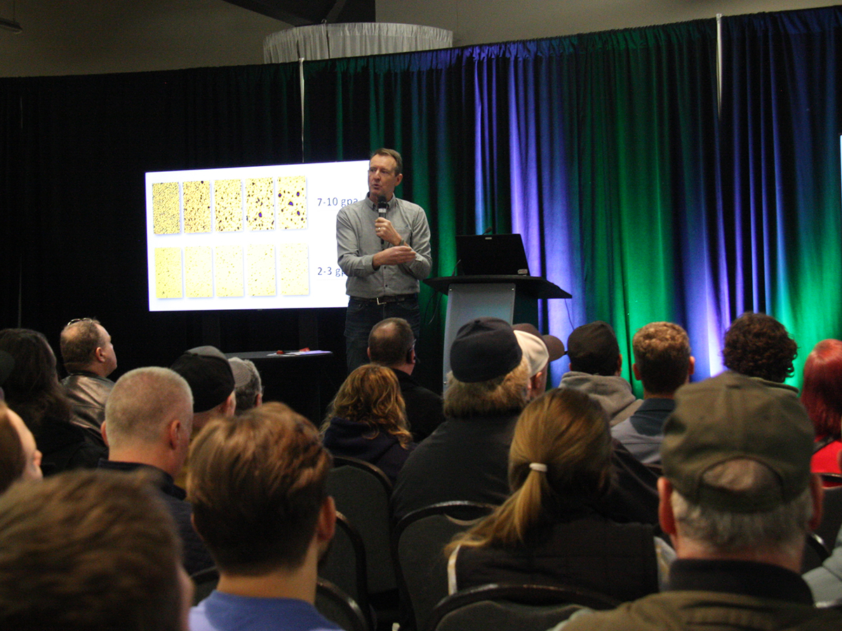 A person stands at a podium, speaking to an audience in a conference room. A presentation slide is displayed in the background.