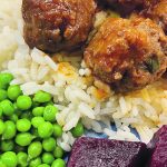 Close-up of a plate with sweet and sour meatballs with steaming rice and a green side of peas or fresh lettuce and some pickled beets.
