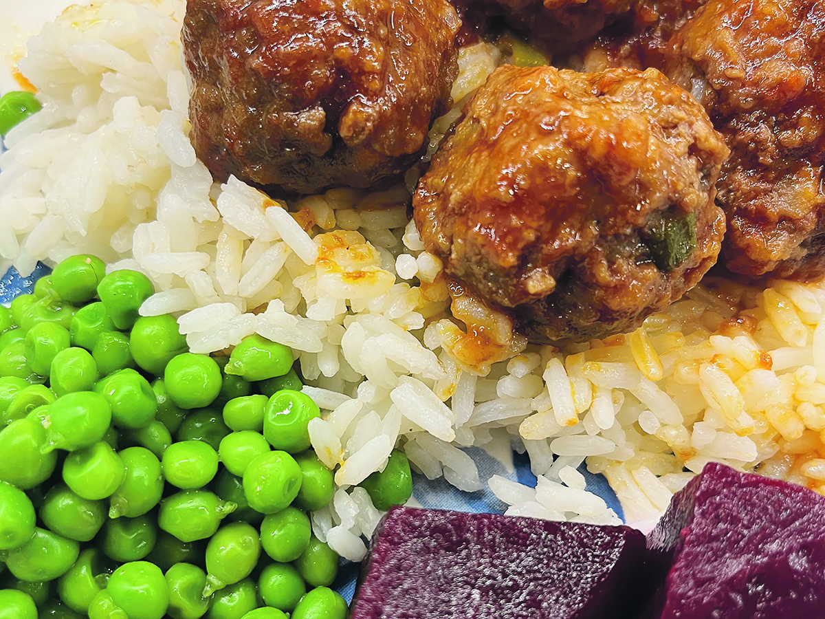Close-up of a plate with sweet and sour meatballs with steaming rice and a green side of peas or fresh lettuce and some pickled beets.