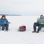 A couple bundled in winter clothing sit outdoors, holding thermoses, in folding chairs in snow in the middle of a lake with a wood fire burning in a round metal stove between them.