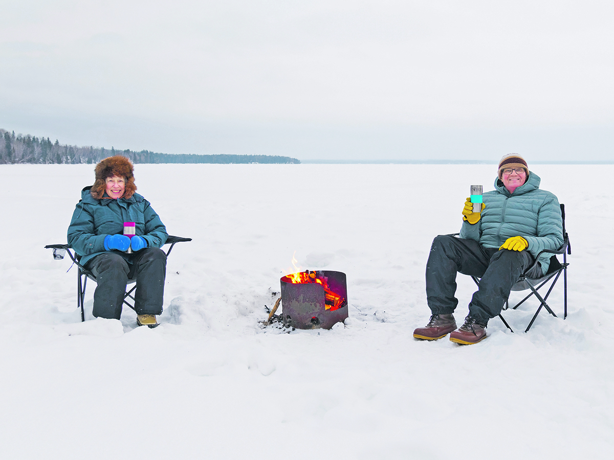 A couple bundled in winter clothing sit outdoors, holding thermoses, in folding chairs in snow in the middle of a lake with a wood fire burning in a round metal stove between them.