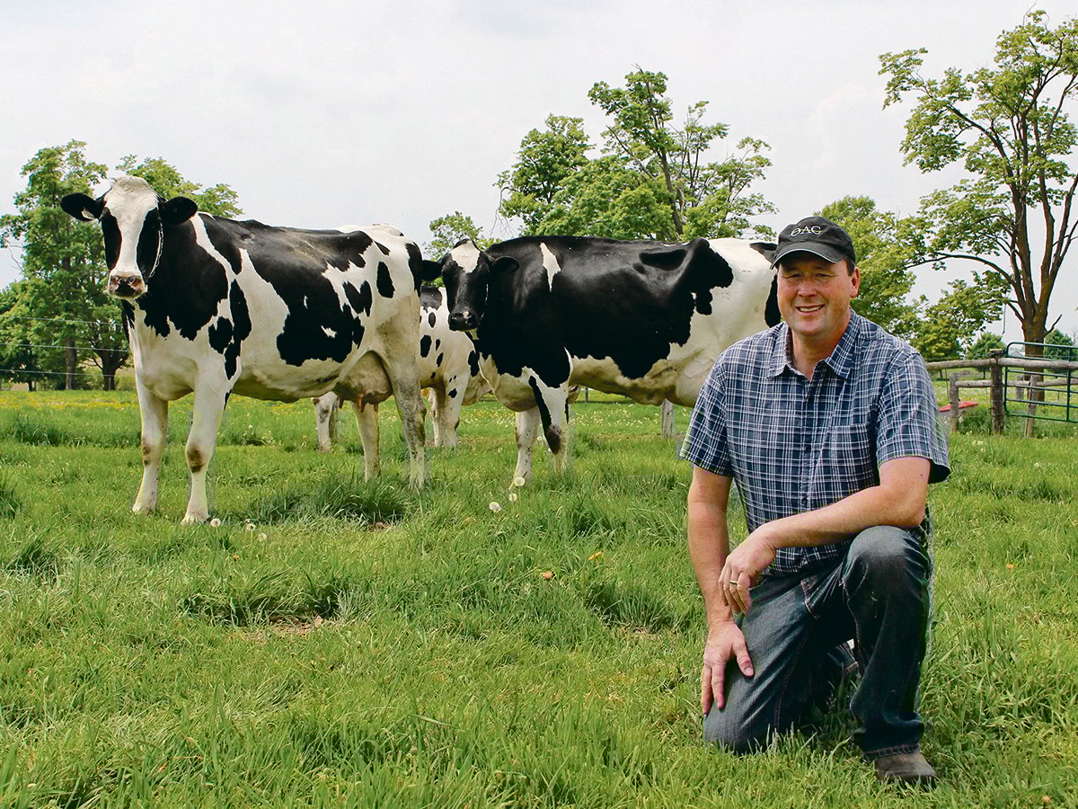 Farmer Tim May kneels in some lush, green grass with some dairy cattle behind him.