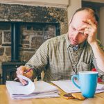 A man sitting at a kitchen table leafing through a large pile of paperwork, obviously frustrated.