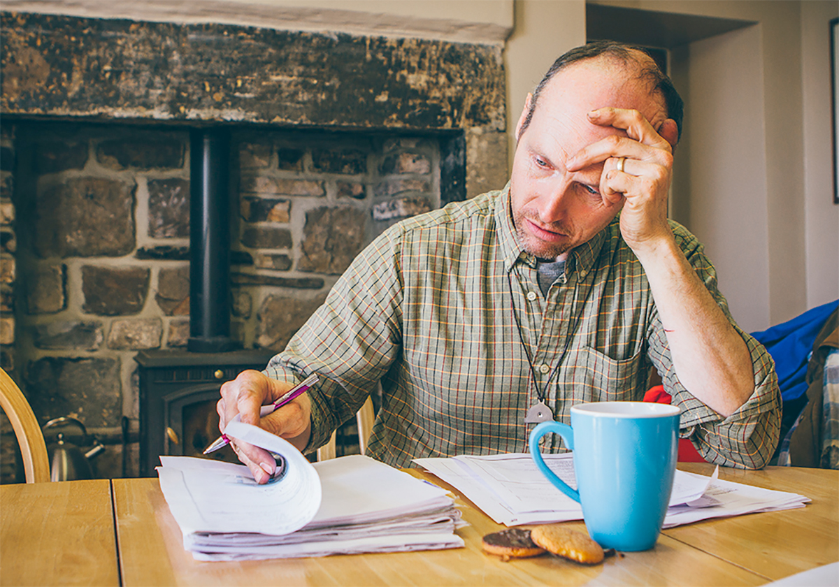 A man sitting at a kitchen table leafing through a large pile of paperwork, obviously frustrated.