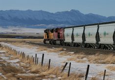 Two train locomotives pull Canpotex cars loaded with potash westward toward the mountains in the distance.