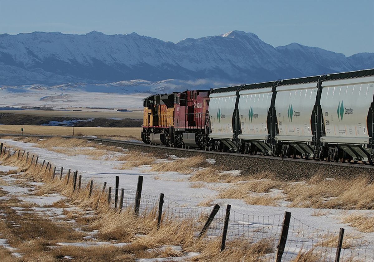 Two train locomotives pull Canpotex cars loaded with potash westward toward the mountains in the distance.