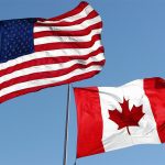 American and Canadian flags fly side-by-side against a clear blue sky background.