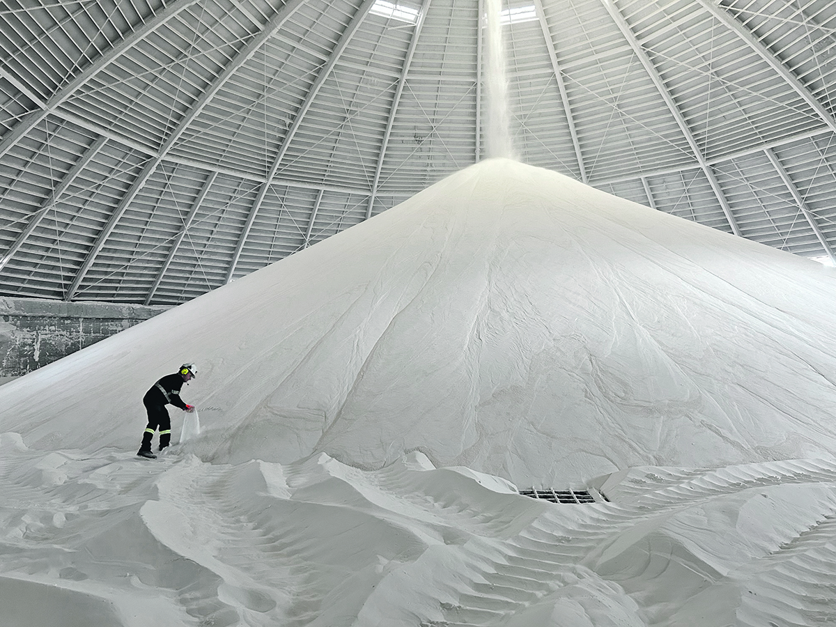 A worker wearing blue safety overalls, a hard hat and hearing protection collects a sample of potassium fertilizer from a huge pile of the substance inside a mine's dome-shaped storage area.