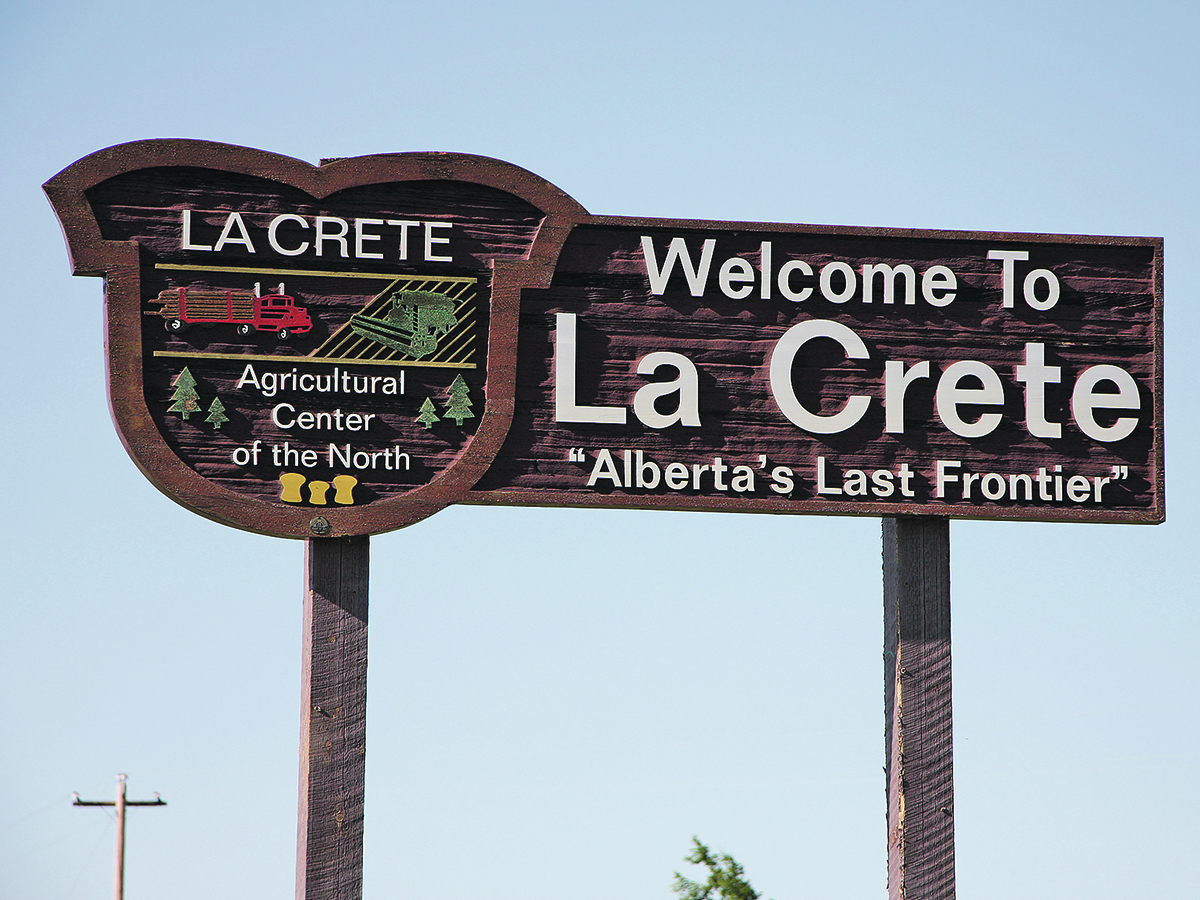 A large rectangular wooden sign like those placed on the edge of town that says, "Welcome to La Crete Alberta's last frontier." The left side of the sign is crest-shaped and has images of a logging truck, a combine, pine trees and wheat sheaves on it and reads, "La Crete - Agricultural center of the north."