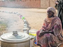 A woman sits as she watches over a large steel pot with a lid as steam rises and a long line of pails and containers of varying colours and sizes sit on the ground snaking off into the distance.