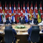 Prime minister Justin Trudeau sits at a large round table with provincial premiers in front of a row of alternating provincial and Canadian flags along the wall in the background.