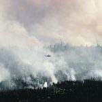 In the distance, a helicopter flies over the smoke rising from a huge forest fire.