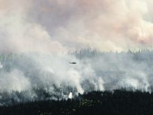 In the distance, a helicopter flies over the smoke rising from a huge forest fire.